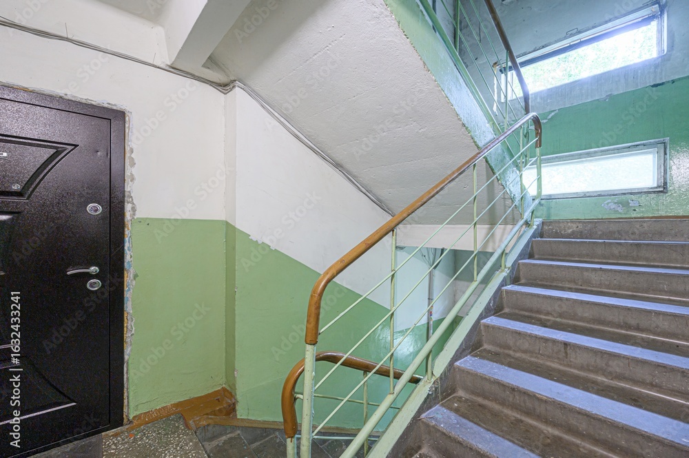 Obraz premium stairwell with concrete steps, light green walls, and a dark door. A metal railing with a wooden handle runs along the stairs