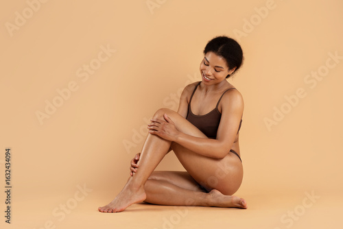 Εκτύπωση καμβά Smiling black woman in underwear sitting cross legged on beige background and looking down in a soft, peaceful pose