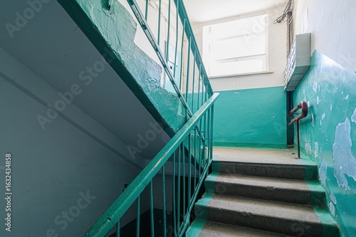 stairwell with green handrails and blue walls, showing signs of age and wear. A window is visible at the landing