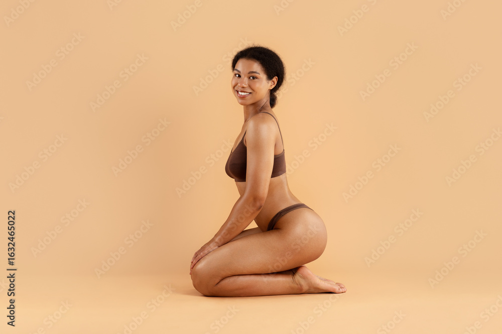 Obraz premium Side view of smiling woman sitting on knees in brown underwear against beige background. Soft lighting and confident pose for wellness, beauty, and comfort concepts.