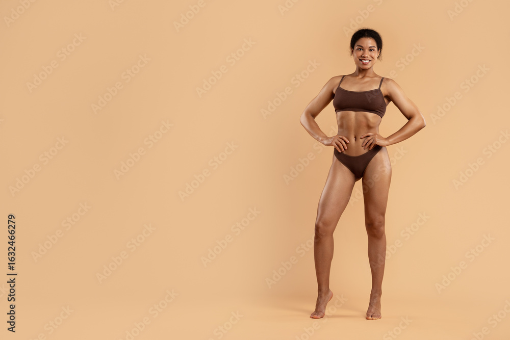 Fototapeta premium Fit African American woman in brown underwear standing with hands on waist, smiling proudly. Beige backdrop and full body shot emphasize wellness and body positivity, copy space