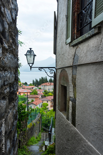 An old street lamp in a gap between old buildings overlooking houses with tiled roofs and Como Lake, Lombardia, Italy