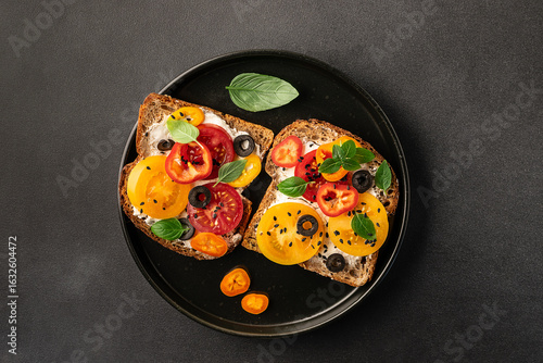 Bright homemade sandwiches with full grain toasts, cream cheese, juicy tomatoes, bell pepper, olives and aromatic herbs on ceramic black plate top view. Healthy tasty breakfast idea.