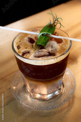 Close-up of a refreshing iced espresso tonic served in a clear glass with ice, garnished with a lime peel and rosemary sprig on a wooden table