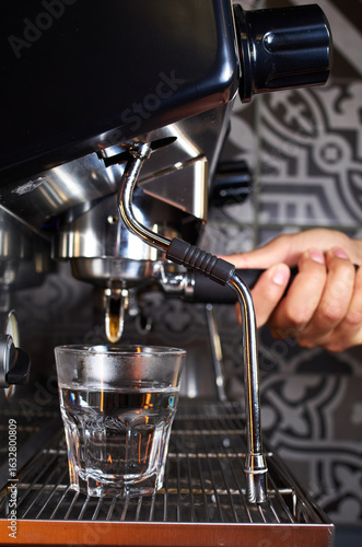 Side view of a barista operating a professional espresso machine, preparing a drink with a transparent glass placed under the group head. The machine's steam wand and the barista’s hand are visible