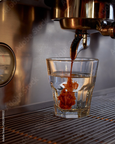 Close-up of a professional espresso machine pouring dark espresso into a transparent glass filled with water. The vivid coffee swirl creates a dramatic ink-like effect inside the glass.