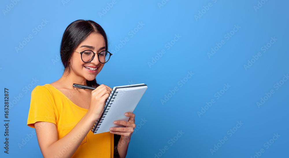 Fototapeta premium Checklist. Portrait of intelligent indian female student wearing glasses, taking notes in copybook on purple studio background. Smart young woman studying, preparing for exam, writing in notebook