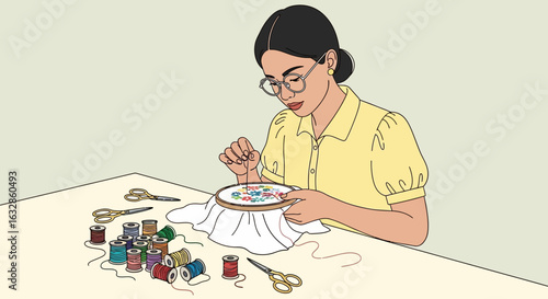 A focused woman with glasses sits at a table, diligently embroidering a colorful pattern.