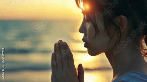Woman is shown in profile, with her hands together in prayerful gesture, set against blurred backdrop sunset over ocean. warm light enhances tranquil, reverent atmosphere.