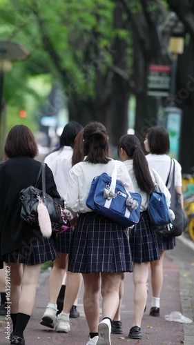 Slow-motion. Japanese high school girls in uniform walk side by side along their school route, seen from behind, with trees and buildings in harmony. Spring. Shanghai's Pudong New District. China.