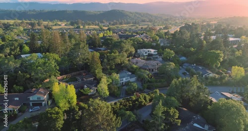 Aerial view of homes nestled among lush trees in Menlo Park, California, USA, residential neighborhood. The sun sets over the hills, casting a warm glow.