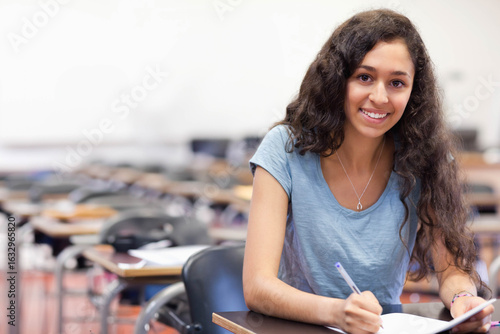 Teenage female student writing in notebook at wooden desk among classroom rows with pen, copy space Free Download Teenage female student writing in notebook at wooden desk among classroom rows with pen, copy space - GFXLoad