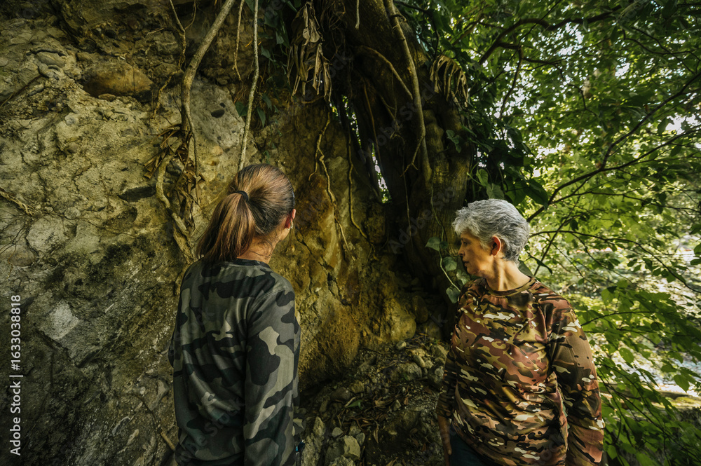 Fototapeta premium Mother and daughter dressed in hiking clothes, walking next to a huge tree that stands on a giant rock in the middle of the rainforest.