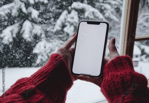 A woman with red knit sleeves holds a smartphone in front of a snowy window, creating a cozy winter vibe during a snowfall. The atmosphere is serene and inviting.