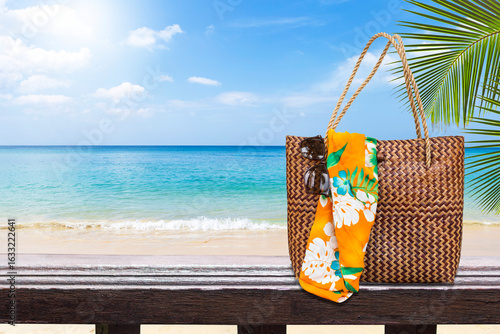 Jute beach bag with sunglasses and colorful cloth on wooden fence over tropical beach, summer outdoor day light, summer season and fashion