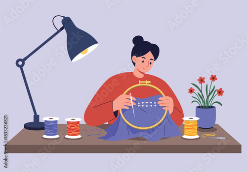 Young woman sitting at a desk and enjoying her needlework hobby. Creative craft of embroidering with a hoop, needle, and thread.