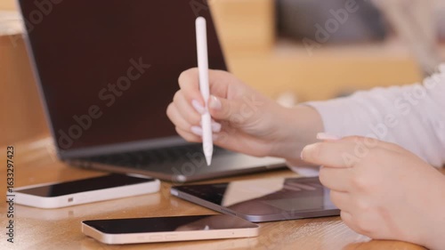 Woman is writing on a tablet with a pen. There are two cell phones on the table, one of which is a flip phone