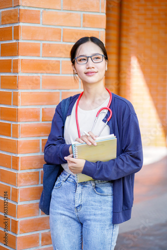 Closeup happiness cheerful Asian young female medical student in casual cloth holding a textbooks and looking out on collage building background. Asian school concept.