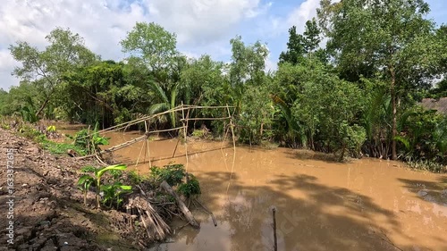 A muddy alluvial canal spanned by a traditional bamboo monkey bridge, surrounded by lush tropical trees in the Mekong Delta, Vietnam.