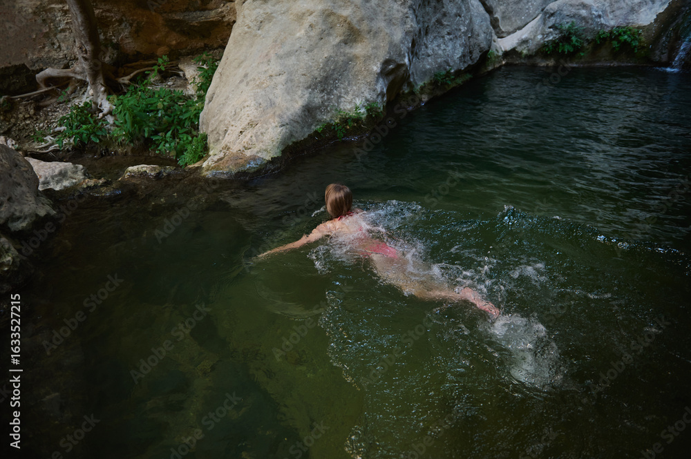 Obraz premium A young woman enjoys a peaceful and serene swim in a natural stream surrounded by rocks and lush greenery, showcasing a moment of relaxation in nature.