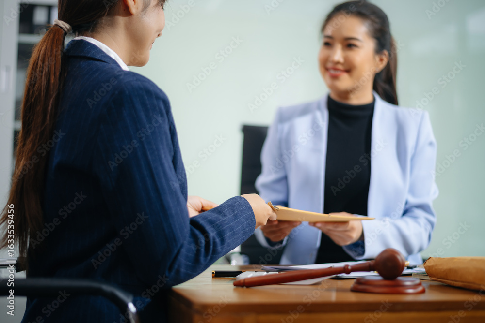 Fototapeta premium Two professional women shake hands over a signed contract at a legal office desk, symbolizing trust, agreement, and partnership.