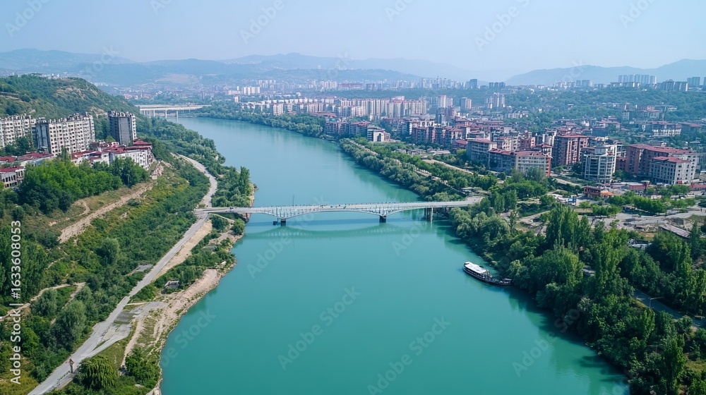 Fototapeta premium High-angle view of a river flowing through a city, with a bridge spanning it. Lush green hills and buildings surround the river