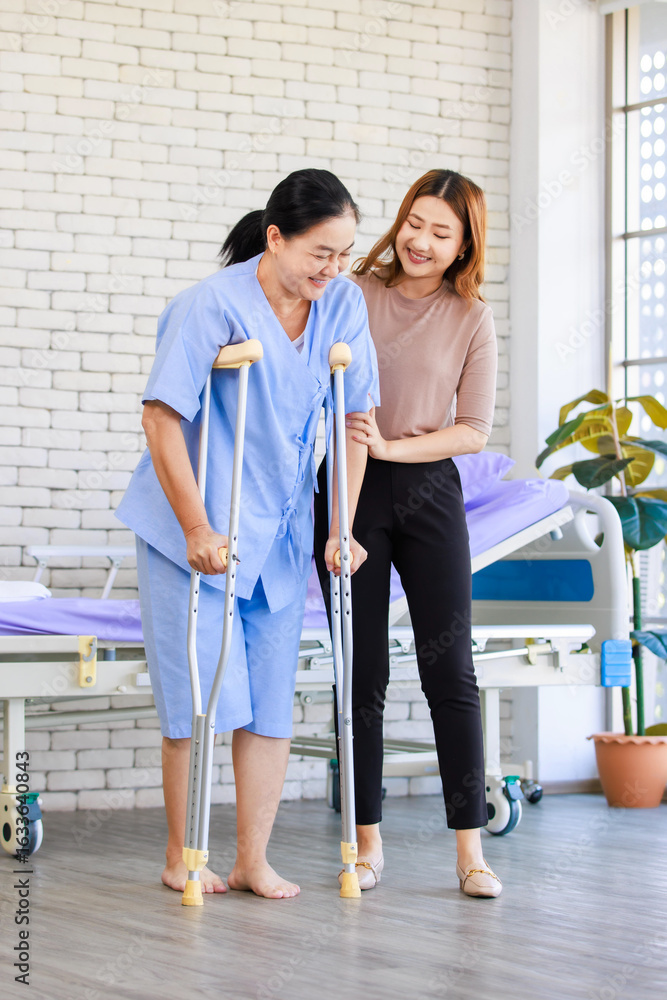 Fototapeta premium Asian young woman teaching walking support crutches walks beside senior female patient in hospital bed room medical recovery caregiver specialist wellness healthcare treatment rehabilitation support.