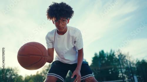 Cute teen boy with curly hair and glasses playing streetball, portrait. Teenager boy training outdoors in summer, future basketballer or streetballer, move to win in championship, hobby and sport