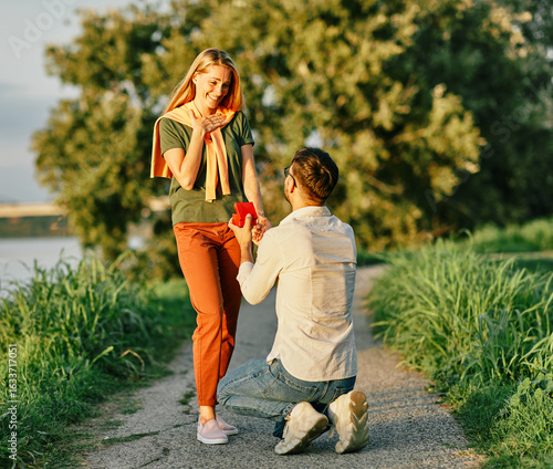 Cheerful handsome man proposing to his girlfriend. Portrait of a young happy couple having fun outdoors in nature during sunset. Girlfriend and boyfriend bonding, love concept during summer.
