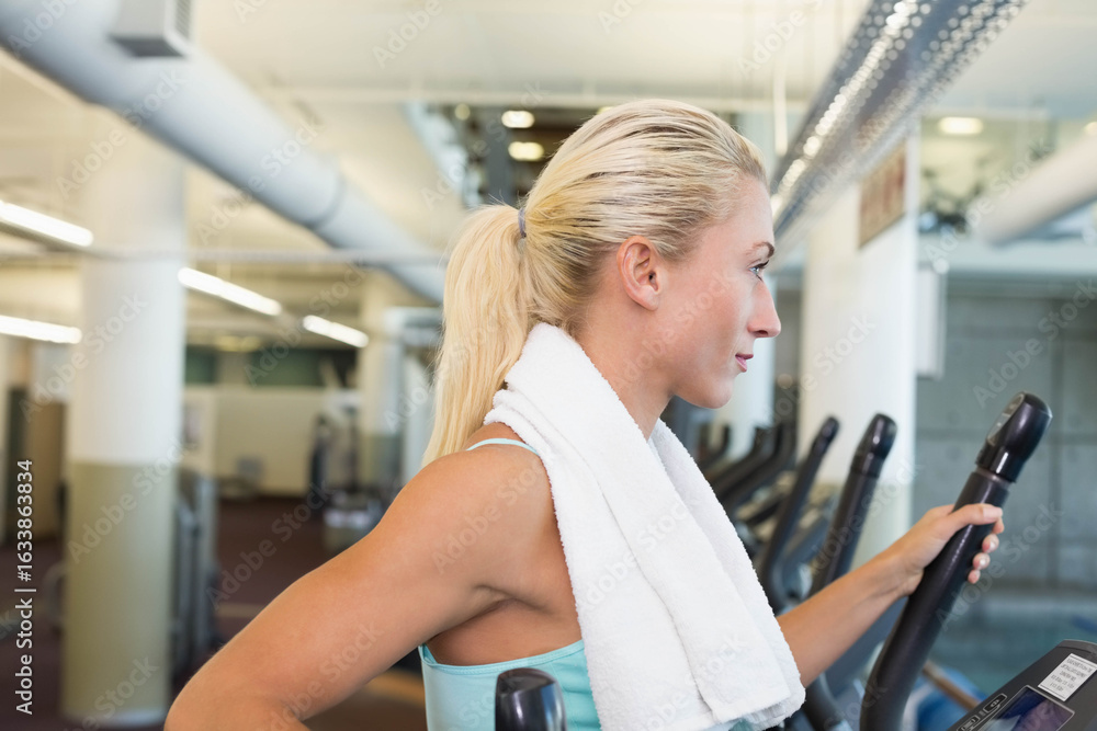 Obraz premium Woman in turquoise tank top wearing gym towel exercising on elliptical machine by ducts, copy space