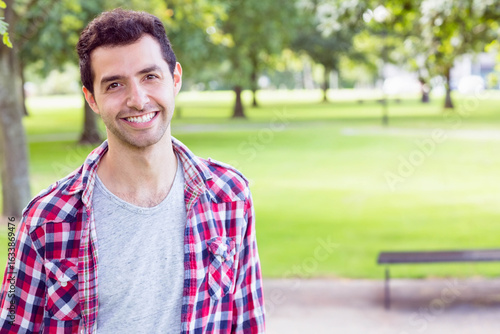 Standing in urban park wearing plaid shirt by wooden bench and paved path, copy space