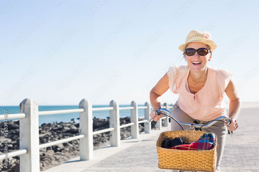Fototapeta premium Woman riding bicycle with basket on seaside promenade by rocky shore, holding blanket and camera