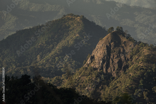 Misty Mountain Ridge with Sunlit Forest at the Summit. Layered green mountains with dramatic sunlight highlighting the forested ridge under a cloudy sky.