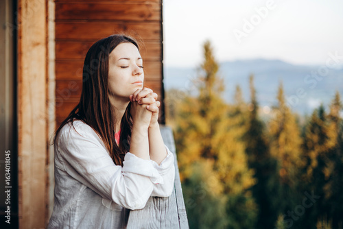 Young woman praying on balcony with mountain view in soft morning light.