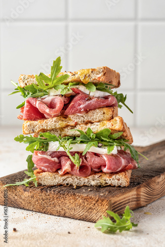 Grilled ham, mozzarella and arugula sandwich on wooden cutting board on white tile kitchen background close up. Delicious breakfast or snack