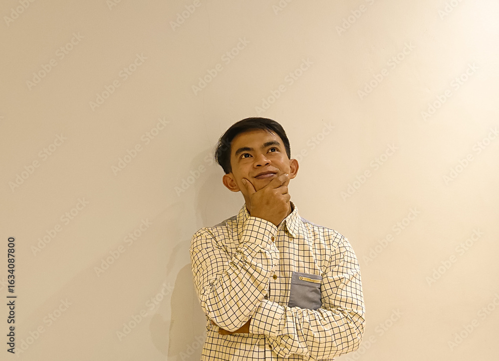Fototapeta premium A young Asian man in a checkered shirt standing against a plain beige wall with a thoughtful expression