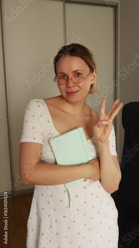 A smiling girl in a light dress and glasses holds a notebook, looks at the camera, and counts with her fingers one, two, three.