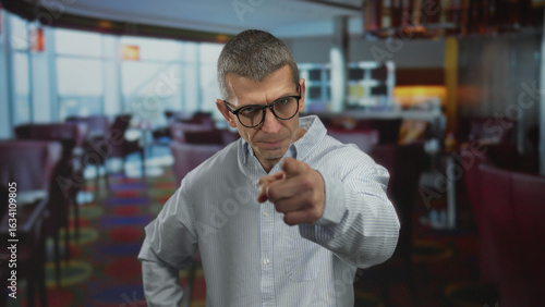 Bilde på lerret Man pointing indoors at a colorful restaurant setting, wearing glasses and a striped shirt, expressing accusation or discipline, with seating and decor blurred in the background