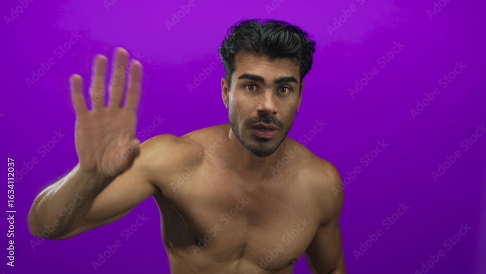 Fototapeta premium Young hispanic man with muscular bare chest points fingers downward in purple studio; invitation.