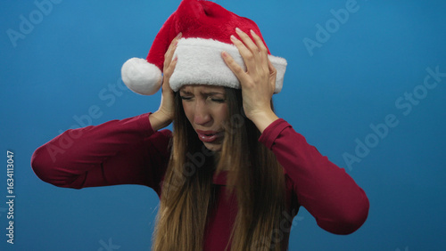 Quadro su tela Young woman in red santa hat pressing hands to head and grimacing in blue studio; holiday stress tension