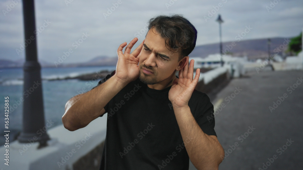 Fototapeta premium Young man standing on a seaside promenade, listening intently with focus against a backdrop of sea and distant hills, embodying curiosity and contemplation of nature's sounds.