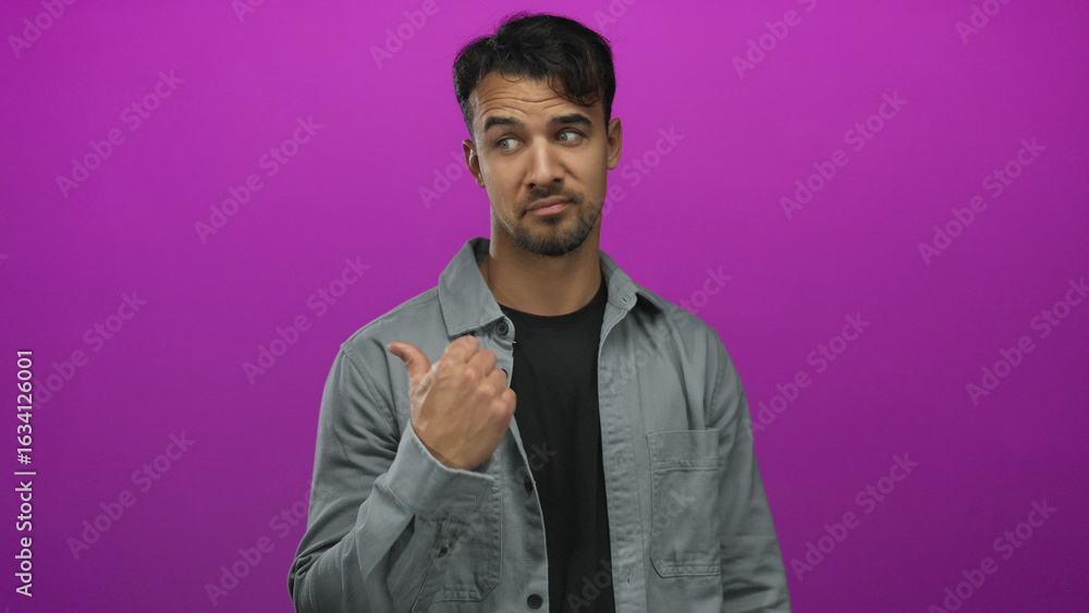 Fototapeta premium Young hispanic man in gray jacket points thumb sideways against vibrant pink background, suggesting indecision or inquiry with a confident expression.