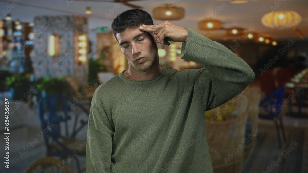 Fototapeta premium Young hispanic man in green sweater makes finger gun to head inside brightly lit building; despair isolation.