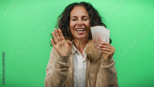 Photography Middle-aged woman holding singapore banknotes smiling and waving against a green wall background, symbolizing wealth and happiness