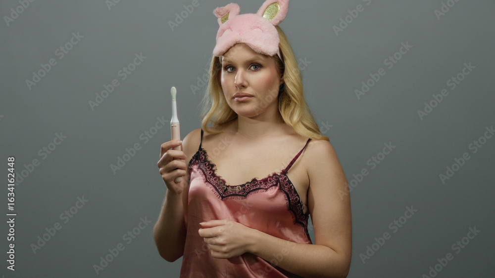 Obraz premium Young blonde woman in sleepwear holding toothbrush against grey background wearing pink sleep mask and examining toothbrush closely