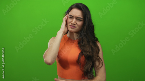Foto Woman in orange top standing upright with long dark hair and hand on cheek experiencing toothache in studio; pain