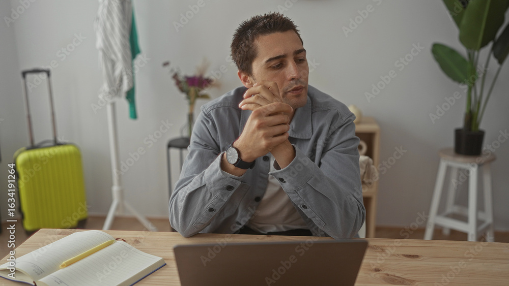 Naklejka premium Young man sitting indoors with hands clasped at wooden desk laptop open behind, thoughtful expression, surrounded by a plant, suitcase, notepad in bright interior living room.