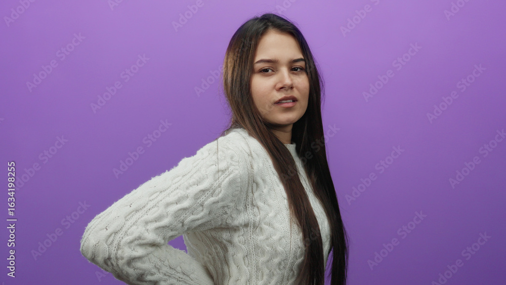 Fototapeta premium Young woman in a white sweater stands with a thoughtful expression against a vibrant purple background, with her hand resting on her lower back as if experiencing discomfort.