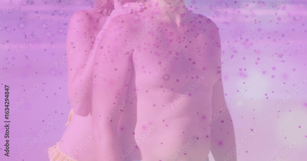 Fototapeta premium Embracing pair wearing dark swim trunks and yellow bikini bottom at beach shoreline, pink overlay