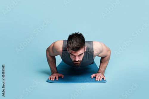 A focused man engages in push-ups on a yoga mat, showcasing strength and determination. The bright blue studio provides an energetic backdrop for his workout routine